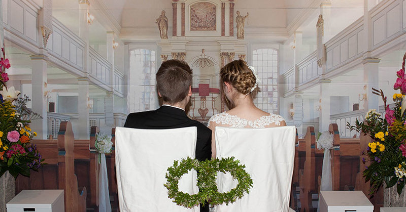 Ein angehendes Hochzeitspaar nimmt auf den Stühlen vor der Bannerwand Platz, die den Altarraum abbildet. 