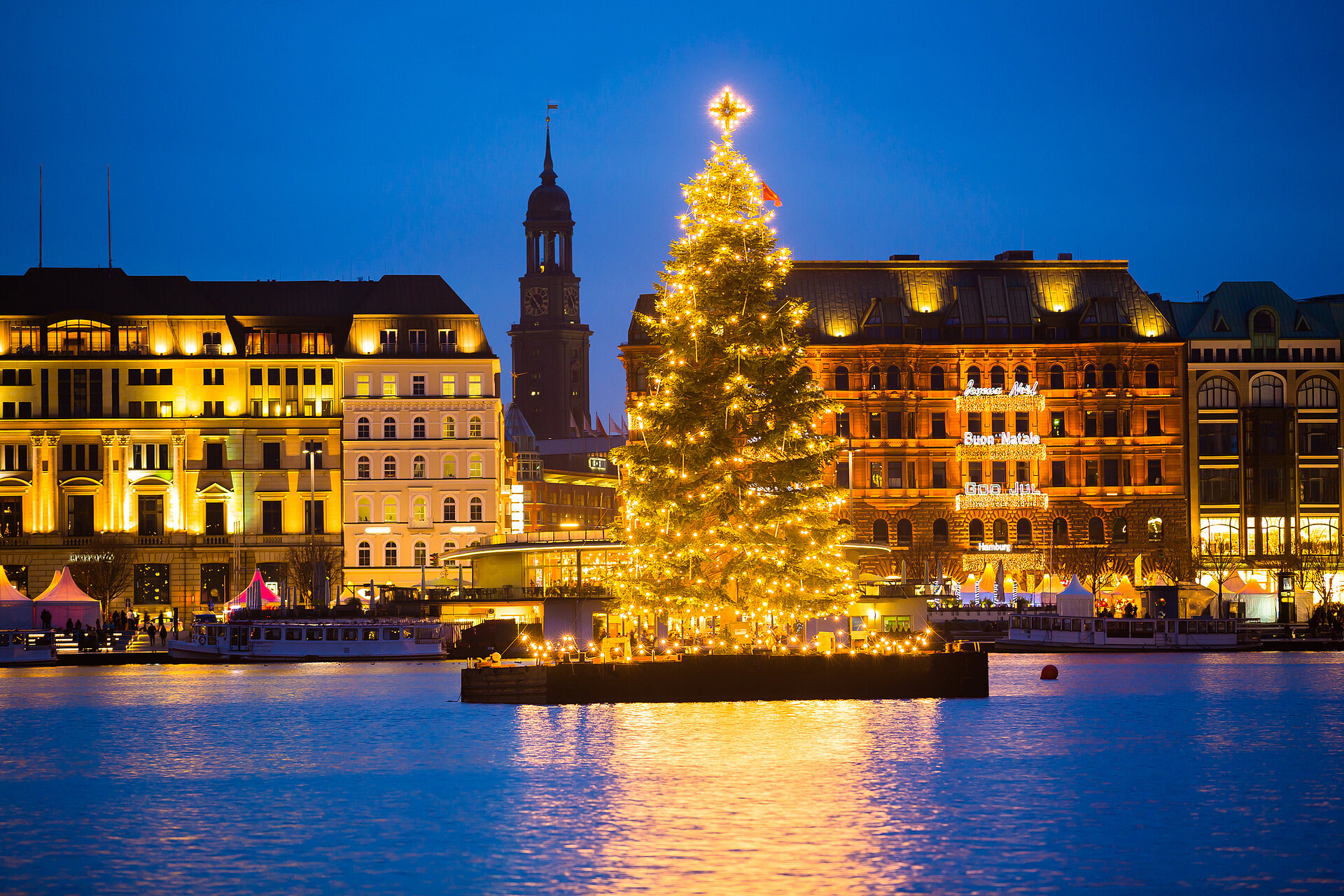 Beleuchtete Weihnachtstanne auf der Hamburger Alster mit Skyline und der Hauptkirche St. Michaelis im Hintergrund.