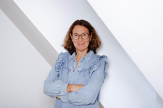 Lächelnde Frau mit runder Brille und hellblauer Jeansbluse, die Arme verschränkt.