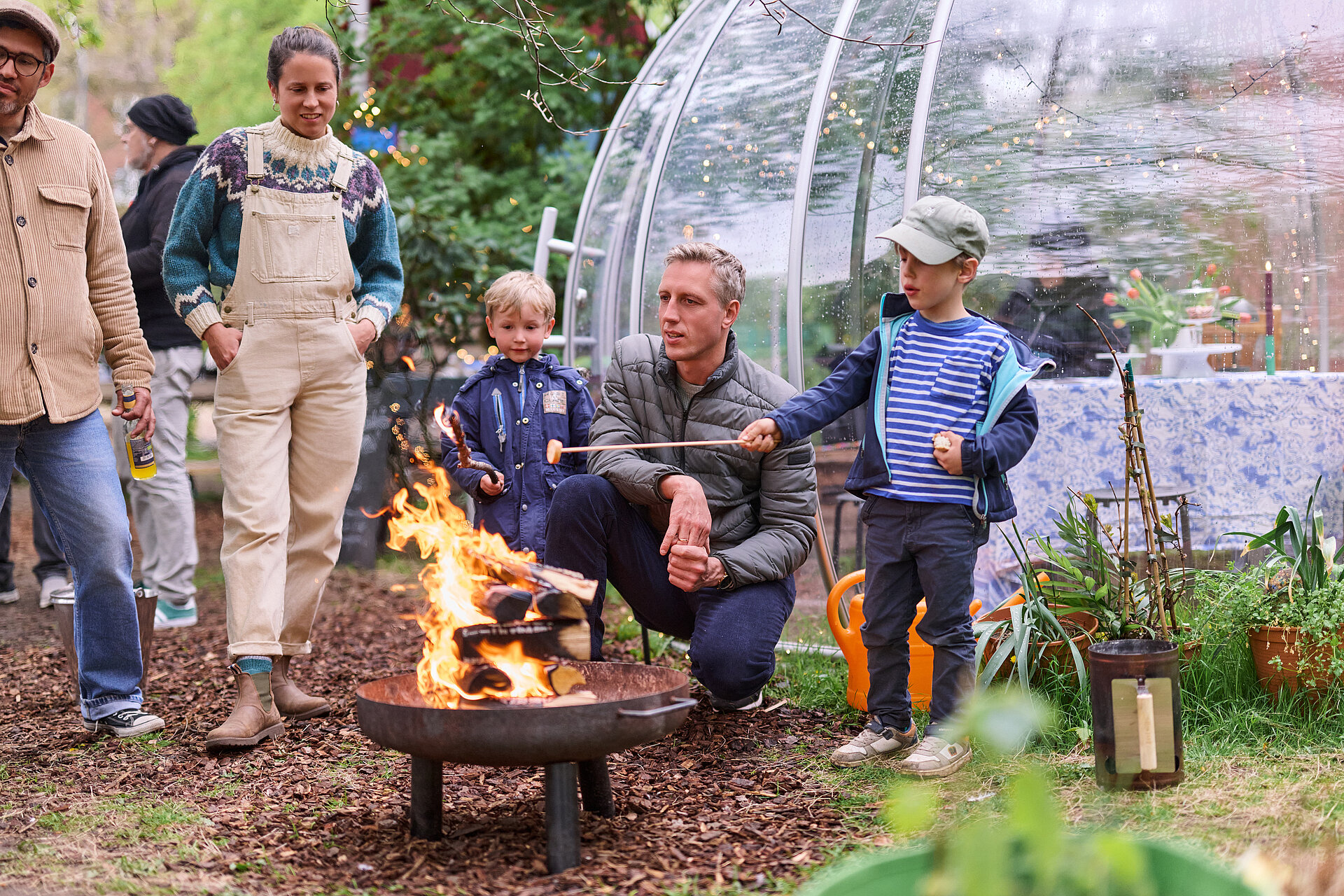 Eine Gruppe von Menschen steht um eine Feuerschale, ein Kind macht Stockbrot
