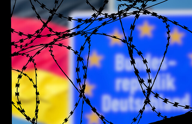 Eine Deutschlandfahne und ein Schild Bundesrepublik Deutschland hinter einem Stacheldraht-Grenzzaun.