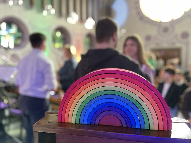 Im Vordergrund steht ein buntes, halbkreisförmiges Holzspielzeug in Regenbogenfarben. Im Hintergrund sind mehrere Menschen in einem Raum mit runden Fenstern und buntem Licht verschwommen zu sehen, was auf eine Veranstaltung oder ein Treffen in freundlicher Atmosphäre hindeutet.