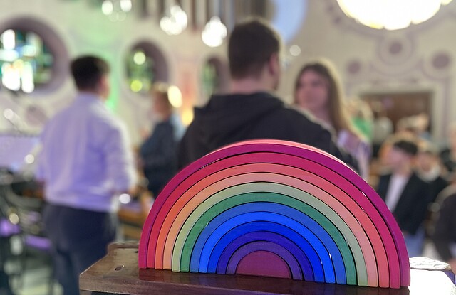 Im Vordergrund steht ein buntes, halbkreisförmiges Holzspielzeug in Regenbogenfarben. Im Hintergrund sind mehrere Menschen in einem Raum mit runden Fenstern und buntem Licht verschwommen zu sehen, was auf eine Veranstaltung oder ein Treffen in freundlicher Atmosphäre hindeutet.