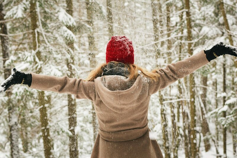 Eine junge Frau im beigen Mantel und einer roten Jacke steht mit ausgebreiteten Armen inmitten eines verschneiten Waldes.