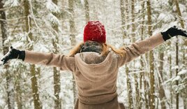 Eine junge Frau im beigen Mantel und einer roten Jacke steht mit ausgebreiteten Armen inmitten eines verschneiten Waldes. - Copyright: Tim Gouw; Pexels